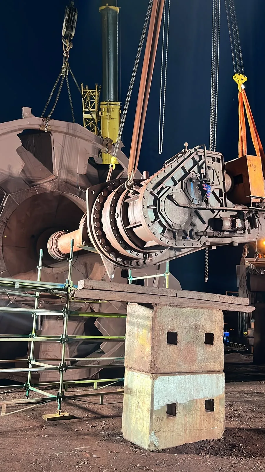 Heavy industrial machinery suspended from slings and chains beside a large wheel assembly.